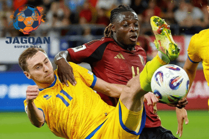 Dua pemain dari tim nasional berbeda berebut bola dalam duel sengit pada pertandingan sepak bola internasional, dengan logo Jagoan Super Bola di sudut kiri gambar.