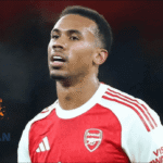 Seorang pemain Arsenal mengenakan jersey merah-putih sedang berada di lapangan saat pertandingan, dengan logo Jagoan Super Bola di bagian kiri gambar.