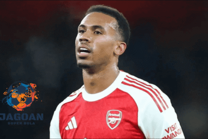 Seorang pemain Arsenal mengenakan jersey merah-putih sedang berada di lapangan saat pertandingan, dengan logo Jagoan Super Bola di bagian kiri gambar.