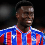 Seorang pemain Crystal Palace mengenakan jersey merah-biru sedang tersenyum di lapangan dalam pertandingan sepak bola.