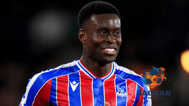 Seorang pemain Crystal Palace mengenakan jersey merah-biru sedang tersenyum di lapangan dalam pertandingan sepak bola.