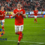 Pemain sepak bola Austria melakukan selebrasi gol di lapangan saat pertandingan kualifikasi Euro, mengenakan jersey merah dan putih dengan sorak penonton di belakangnya.