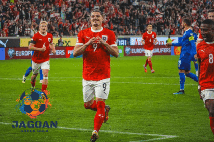 Pemain sepak bola Austria melakukan selebrasi gol di lapangan saat pertandingan kualifikasi Euro, mengenakan jersey merah dan putih dengan sorak penonton di belakangnya.