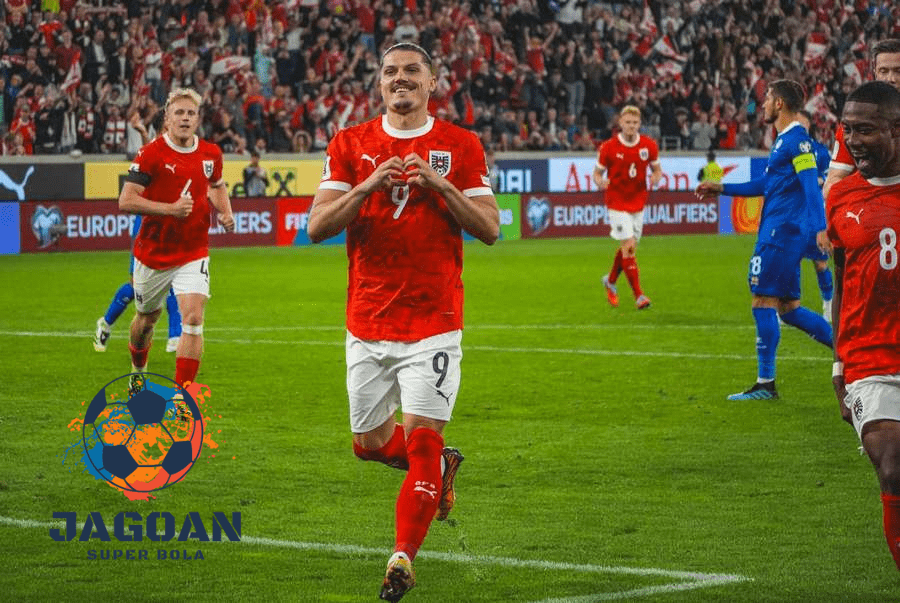 Pemain sepak bola Austria melakukan selebrasi gol di lapangan saat pertandingan kualifikasi Euro, mengenakan jersey merah dan putih dengan sorak penonton di belakangnya.