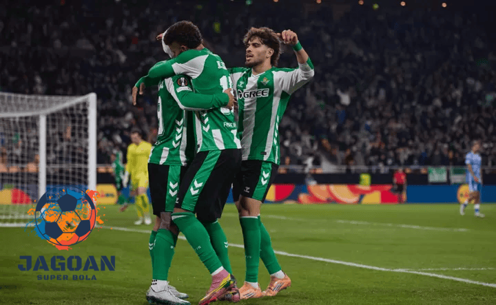 Pemain sepak bola Real Betis merayakan gol bersama di tengah pertandingan dengan logo Jagoan Super Bola pada sudut gambar.