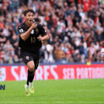 Pemain tim sepak bola merayakan gol di stadion penuh penonton, logo JagoanSuperBola di pojok kiri bawah