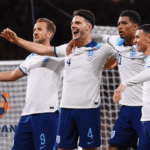 Pemain timnas Inggris merayakan gol bersama di lapangan sepak bola saat laga internasional, mengenakan jersey putih biru dengan ekspresi gembira.