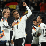 Para pemain tim nasional Jerman merayakan gol di lapangan dengan mengenakan jersey putih bergaris warna bendera Jerman.