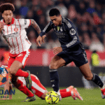 Dua pemain berebut bola dalam pertandingan sepak bola dengan intensitas tinggi di lapangan dengan logo Jagoan Super Bola di sudut gambar.