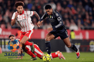 Dua pemain berebut bola dalam pertandingan sepak bola dengan intensitas tinggi di lapangan dengan logo Jagoan Super Bola di sudut gambar.