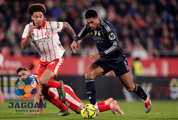 Dua pemain berebut bola dalam pertandingan sepak bola dengan intensitas tinggi di lapangan dengan logo Jagoan Super Bola di sudut gambar.