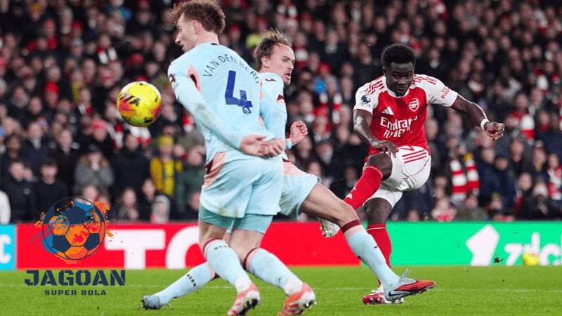 Pemain Arsenal menendang bola ke arah gawang melawan dua bek Brentford dalam pertandingan sepak bola, dengan logo Jagoan Super Bola di sudut kiri gambar.