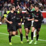 Pemain Bayer Leverkusen mengenakan jersey hitam dengan sponsor Barmenia merayakan gol di pertandingan sepak bola kandang dengan latar penonton penuh.