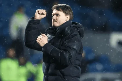 Manajer sepak bola Danny Röhl mengepalkan tangan merayakan kemenangan di bawah guyuran hujan di stadion, merepresentasikan momentum tim.