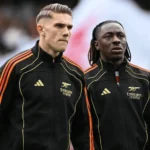 Eberechi Eze dan Viktor Gyökeres mengenakan jersey training hitam bergaris oranye Adidas dengan sponsor Emirates, berhadapan di dalam stadion selama sesi pemanasan atau pertandingan.