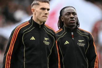 Eberechi Eze dan Viktor Gyökeres mengenakan jersey training hitam bergaris oranye Adidas dengan sponsor Emirates, berhadapan di dalam stadion selama sesi pemanasan atau pertandingan.