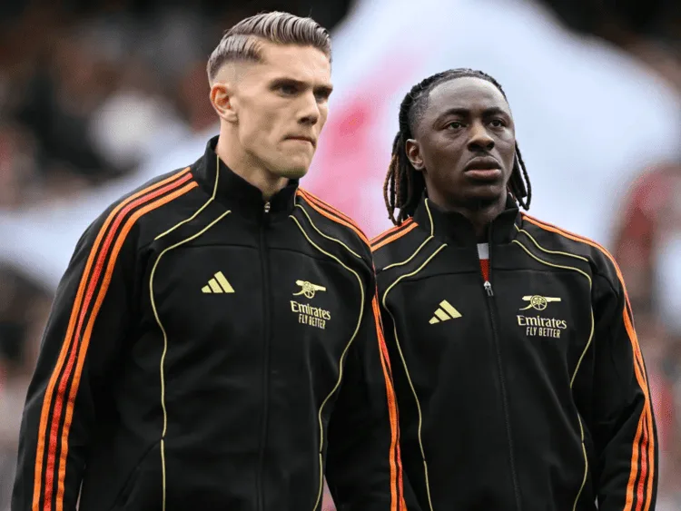Eberechi Eze dan Viktor Gyökeres mengenakan jersey training hitam bergaris oranye Adidas dengan sponsor Emirates, berhadapan di dalam stadion selama sesi pemanasan atau pertandingan.