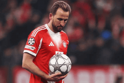 Harry Kane mengenakan seragam merah dengan logo Adidas dan lencana Champions League, menunduk sambil memegang bola bertabur bintang di depan penonton yang blur