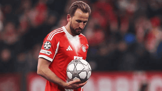 Harry Kane mengenakan seragam merah dengan logo Adidas dan lencana Champions League, menunduk sambil memegang bola bertabur bintang di depan penonton yang blur