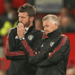 Ole Gunnar Solskjaer, former Manchester United manager, stands alongside bearded assistant coach on Old Trafford touchline, both in red Adidas tracksuits with club crest