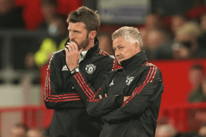 Ole Gunnar Solskjaer, former Manchester United manager, stands alongside bearded assistant coach on Old Trafford touchline, both in red Adidas tracksuits with club crest