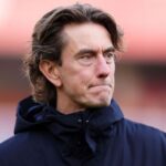 Foto close-up Thomas Frank sebagai manager Tottenham Hotspur dengan ekspresi murung dan serius, mengenakan jaket biru tua berhoodie di pinggir lapangan sepak bola dengan latar belakang pink saat cuaca dingin.