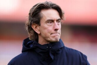 Foto close-up Thomas Frank sebagai manager Tottenham Hotspur dengan ekspresi murung dan serius, mengenakan jaket biru tua berhoodie di pinggir lapangan sepak bola dengan latar belakang pink saat cuaca dingin.