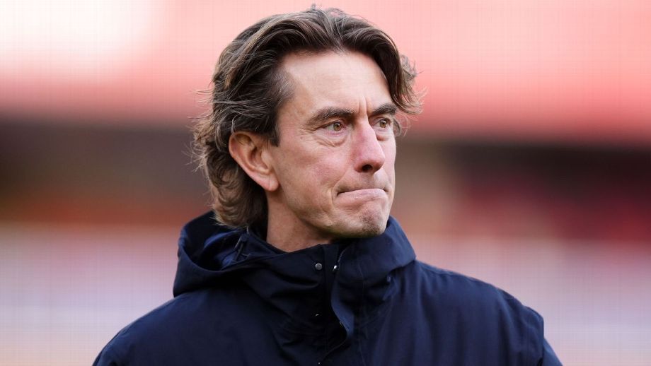 Foto close-up Thomas Frank sebagai manager Tottenham Hotspur dengan ekspresi murung dan serius, mengenakan jaket biru tua berhoodie di pinggir lapangan sepak bola dengan latar belakang pink saat cuaca dingin.