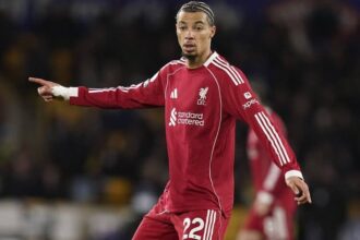 Hugo Ekitike Liverpool FC nomor 22 mengenakan jersey merah Adidas panjang lengan sponsor Standar Chartered, berpose dengan jari telunjuk di lapangan sepak bola - Bola188